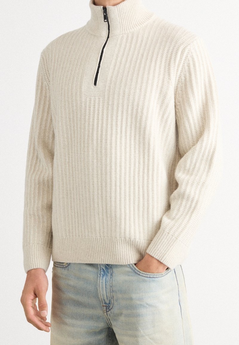 Homme portant un pull en maille côtelée couleur crème avec une fermeture éclair noire au quart et un jean bleu clair, une main dans la poche.