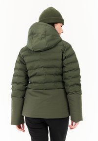 Donkergroene pufferjack met een capuchon, voorzien van horizontale gestikte secties en een getailleerde taille. Het model draagt ook een bijpassende groene gebreide muts.
