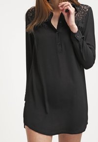 Blusa nera con maniche lunghe, chiusura frontale con bottoni e dettagli in pizzo sulle spalle, realizzata in tessuto leggero con una texture liscia.