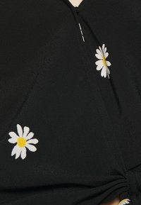 Top nero con scollatura a V, decorato con ricami di margherite bianche e gialle, e con un dettaglio a nodo all'orlo. Tessuto morbido e testurizzato.