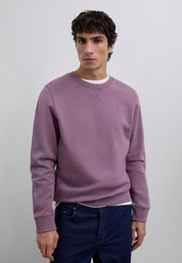 Joven vistiendo un suéter de cuello redondo color malva sobre una camisa blanca y unos jeans azul oscuro, de pie contra un fondo gris claro liso.