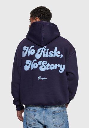 Muž v tmavomodrej mikine s kapucňou s bledomodrým nápisom „No Risk, No Story“ a logom „Dropsize“ na chrbte, v kombinácii so svetlými džínsami.