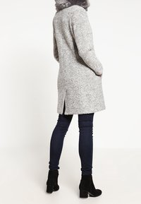 Grijze textuurjas met een nepbontkraag en zijsplitten, gecombineerd met donkere skinny jeans en zwarte enkellaarsjes.