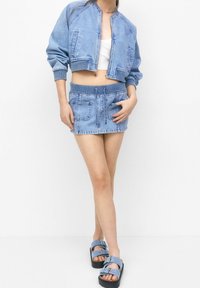 Lichtblauwe denim cropped jack met geribbelde manchetten, gecombineerd met een bijpassende denim minirok met frontzakken en trekkoord. Blauwe sandalen.