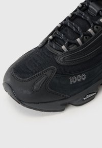 Svart treningssneaker med nettingoverdel, teksturerte gummidetaljer, polstret krage og reflekterende detaljer. Har "1000" merkevarenavn på siden.