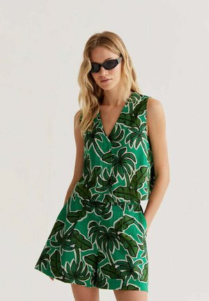 Mujer con top sin mangas verde y pantalones cortos con un gran estampado de hojas de palma en verde oscuro, complementada con gafas de sol negras.