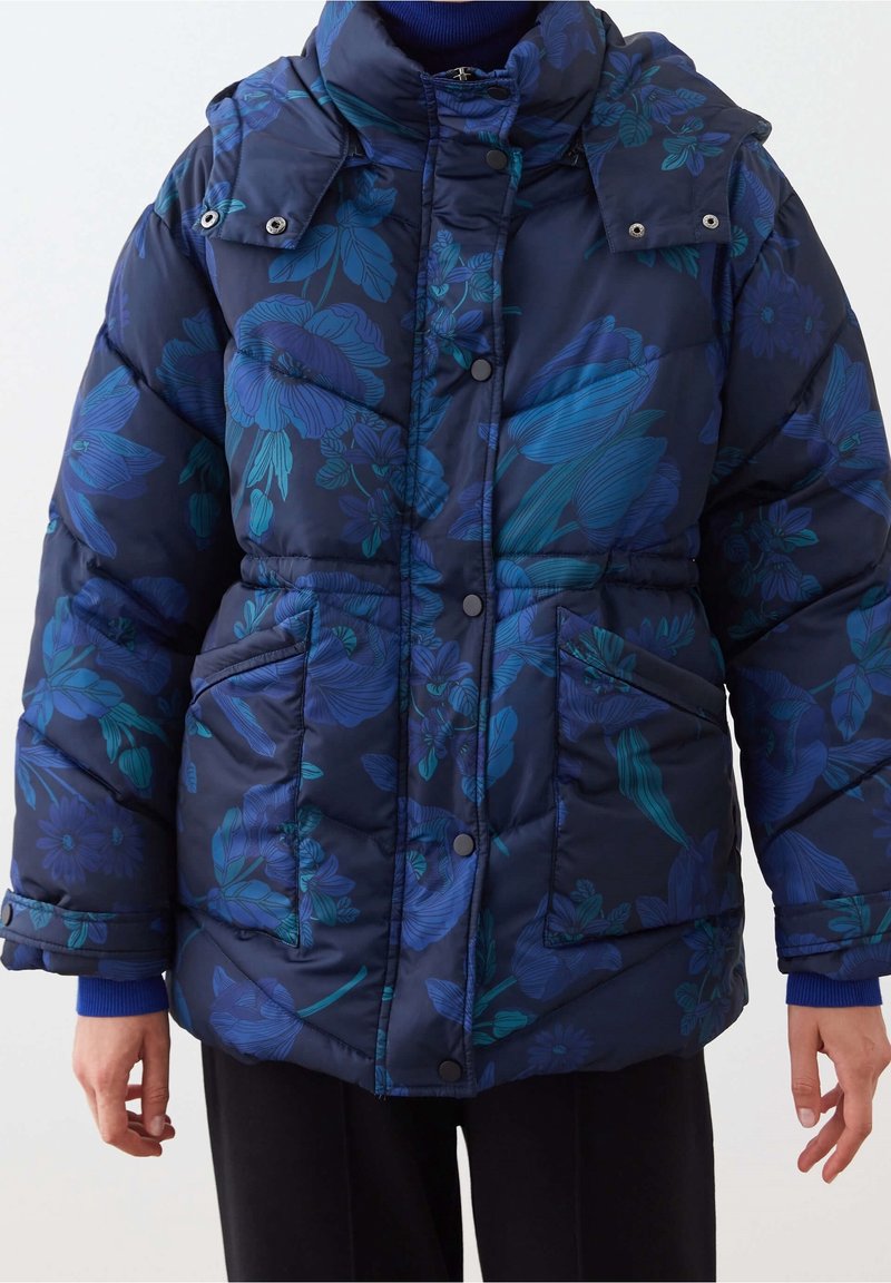 Giacca imbottita navy con motivo floreale in tonalità di teal e blu. Presenta un colletto alto, bottoni a pressione e due tasche frontali.