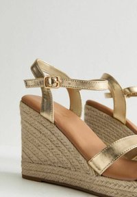 Sandales compensées en métal doré avec une plateforme en jute tressé et une bride autour de la cheville. Texture lisse sur les brides avec un détail de boucle.