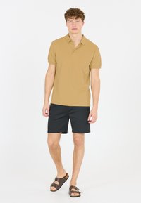 Tan polo shirt med två knappar, i kombination med svarta shorts och bruna sandaler. Tyget verkar slätt och lätt.