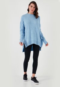 Gaiši zils adīts džemperis ar garām piedurknēm un augstu-zemu apakšmalu, kombinēts ar melniem šauriem džinsiem un melniem sporta apaviem. Gluda tekstūra, ikdienišķa piegriezuma.