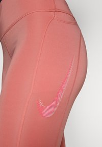 Różowe legginsy sportowe z gładkiego, elastycznego materiału. Posiadają białą i różową paskowaną logo Nike po boku oraz widoczne detale szycia.