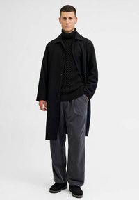 Zwarte lange jas over een zwart textuur turtleneck, gecombineerd met grijze wijde broek en zwarte schoenen. Eenvoudig ontwerp, geen patronen of accenten.
