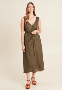 Olivgrünes, ärmelloses Maxikleid mit V-Ausschnitt und Knopfleiste. Verfügt über eine Bindegürtel und eine fließende Textur, kombiniert mit braunen Plateausandalen.