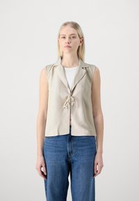 Beige liinapoolne vest, millel on krae, lühikesed varrukad ja sidumisnööp ees, koos siniste kõrge vöökohaga teksastega. Lihtsad, puhtad jooned.
