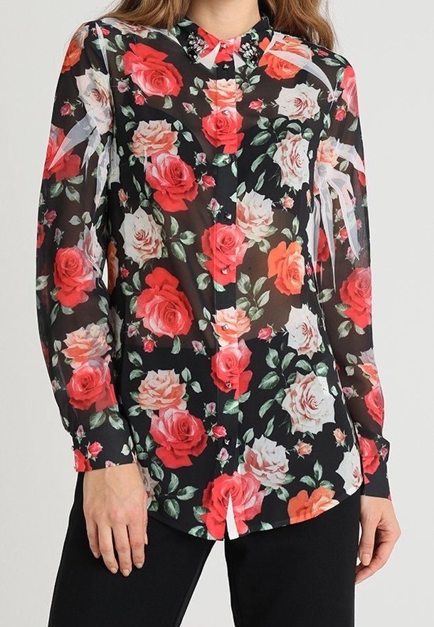 Femme portant un chemisier noir transparent avec un motif floral de roses rouge, rose et blanc éclatant, associé à un pantalon noir.