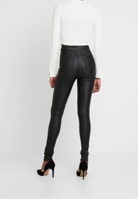 Höga svarta byxor i konstläder med en slät, glänsande yta, som har två bakfickor och är parat med svarta högklackade skor.