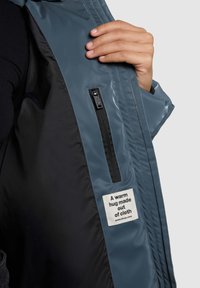 Mėlynos vandeniui atsparios striukės vidus su juodu pamušalu, kuriame yra kišenė su užtrauktuku ir audinio etiketė, kurioje parašyta „Šiltas apkabinimas iš audinio.“