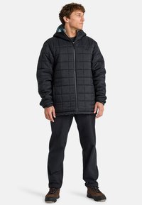 Chaqueta negra acolchada con cierre frontal, diseño acolchado y capucha. Combinada con pantalones negros y botas de senderismo marrones.