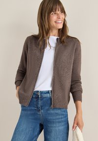 Brun stickad cardigan med ribbade muddar och nederkant, dragkedja framtill, bärs över en vit t-shirt, i kombination med blå jeans. Mjuk textur.
