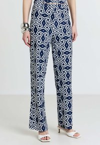 Pantalon bleu marine avec un motif géométrique blanc, présentant un design évasé et arrivant à la cheville. Porté avec des chaussures blanches ouvertes à l'avant et un bracelet en argent.
