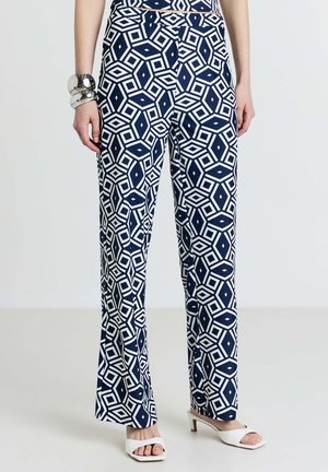 Navy blue pants with a geometric white pattern, featuring a flared design and ankle-length. Worn with white open-toe shoes and a silver bracelet.