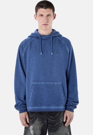 Mladý muž v modré mikině s kapucí, šňůrkami a přední kapsou, v kombinaci s černými potrhanými džíny.