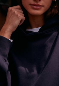Marinbl�å sweatshirt med hög krage, strukturerad tyg och en synlig vit skjorta under. Närbild av hand som greppar kragen.
