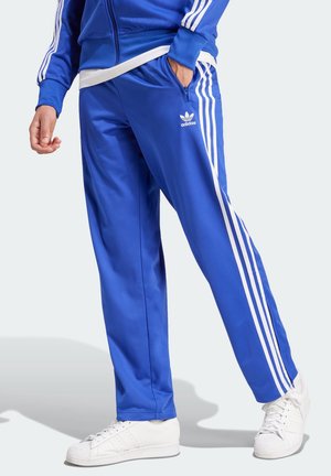 Osoba nosząca niebieskie spodnie dresowe i kurtkę Adidas z białymi paskami, stojąca z jedną ręką w kieszeni i widoczne białe sneakersy.