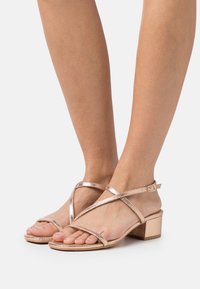 Roséguld klacksandaler med metalliska korsremmar och en texturerad blockklack. Öppen tå-design som framhäver naturliga tår och fötter.
