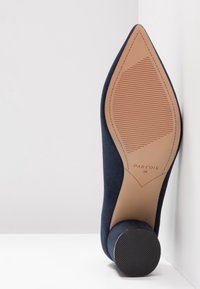 Chaussure bleu marine à bout pointu posée sur son talon noir arrondi, montrant sa semelle marron clair avec une texture antidérapante et la taille 38 indiquée.