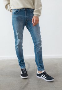 Jeans de denim azul ajustados con parches desgastados, con un diseño clásico de cinco bolsillos. Combinados con zapatillas negras y blancas.