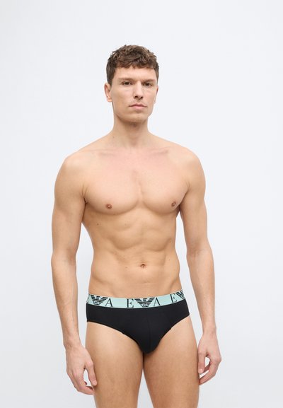 Άνδρας χωρίς πουκάμισο όρθιος, φοράει μαύρα εσώρουχα με ανοιχτό μπλε ζώνη μέσης που φέρει επαναλαμβανόμενο κείμενο «A E» και λογότυπα αετού.