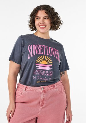 Krøllet-håret kvinde smiler, iført en grå "Sunset Lover" t-shirt med en solnedgangsgrafik og lyserøde cowboybukser med høj talje.