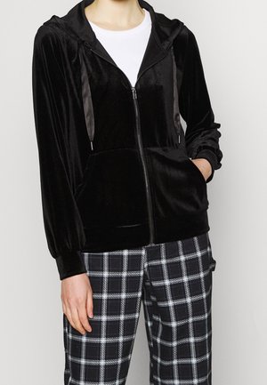 Personne portant un sweat à capuche zippé en velours noir avec cordons et un pantalon à carreaux noir et blanc, main dans la poche du sweat.