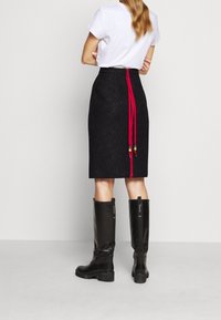 Vrouw die een zwarte kokerrok van kant tot op de knie draagt met een rode rits en franjes aan de achterkant, een wit shirt met korte mouwen en zwarte knie-laarzen.