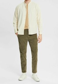 Camicia button-up beige in tessuto leggero, indossata sopra una t-shirt color sabbia; abbinata a pantaloni in corduroy verde e sneakers bianche.