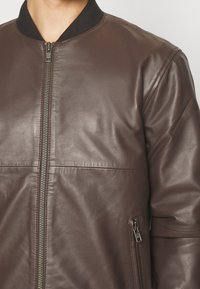 Giacca bomber in pelle marrone con collo a coste nero, cerniera frontale e tasche laterali. Presenta una texture liscia e una vestibilità su misura.