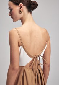 Beige mekko, jossa on tiukka yläosa ja avoin selkä, ohuilla olkaimilla ja solmittavalla kiinnityksellä. Sileä kangas ja laskostettu hame. Kultaista renkaankorviksia.