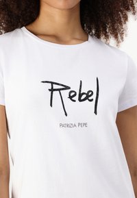 T-shirt bianca a maniche corte con la scritta "Rebel" in bold nera e "Patrizia Pepe" in testo più piccolo sottostante. Materiale in cotone morbido.