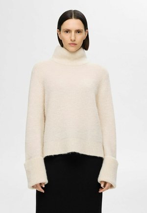 Light beige turtleneck sweater with a chunky, textured knit. Features wide ribbed cuffs and an oversized, boxy fit, layered over a black garment.