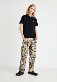 T-shirt nera a maniche corte con logo sul petto, abbinata a pantaloni cargo in camouflage a vestibilità ampia nei colori sabbia, verde e nero, indossati con scarpe nere.