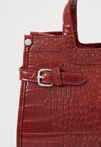 Sac à main en cuir texturé rouge avec un motif crocodile, détails en boucle argentée et poignées robustes. Design compact avec une forme structurée.