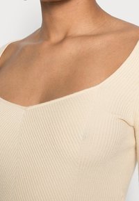 Pehmeäbeige ribbineulottu paita, jossa leveä pyöreä pääntie ja vartalonmyötäinen siluetti hienovaraisilla pystysuorilla viivoilla ja saumattomilla reunoilla.