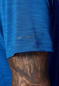 Primo piano di un braccio tatuato parzialmente coperto da una camicia a maniche corte blu testurizzata con un piccolo dettaglio a striscia riflettente.
