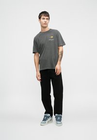 Graues, kurzärmeliges T-Shirt mit dem Schriftzug "AUSTIN, TEXAS" in Orange. Kombiniert mit schwarzen Hosen und hellblauen High-Top-Sneakern.