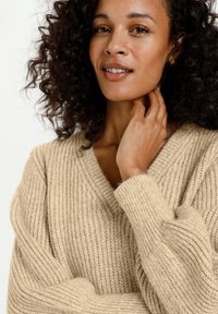 Pull beige en tricot avec un col en V, une texture côtelée et des manches volumineuses. Le modèle touche délicatement son cou tout en souriant.