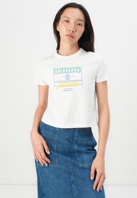 T-shirt blanc court avec un graphique "tommy jeans" en vert et jaune ; associé à une jupe en denim foncé avec des poches et des coutures visibles.