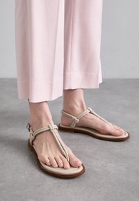 Sandales plates beiges avec un design à lanière en T, présentant des détails noués et une semelle marron. Portées avec un pantalon large rose clair.