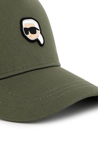 Olíva zöld sapka ívelt karimával, fekete és fehér karikatúra folttal egy napszemüveget viselő arccal. Strukturált dizájn, varrási részletekkel.