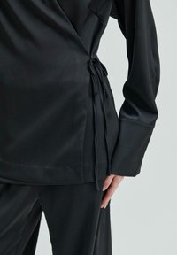 Černý vrchní top z hladkého materiálu má detail v podobě bočního zavázání a dlouhé rukávy, s volným střihem a minimalistickým designem.